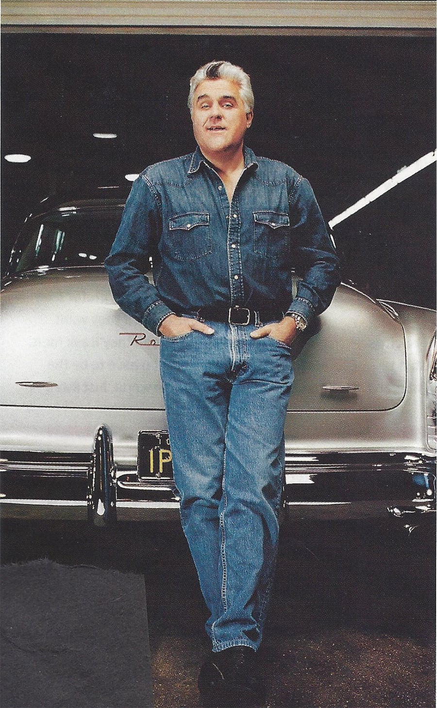 A man with gray hair in a denim shirt and jeans stands casually with hands in his pockets, leaning against a vintage silver car in a garage.