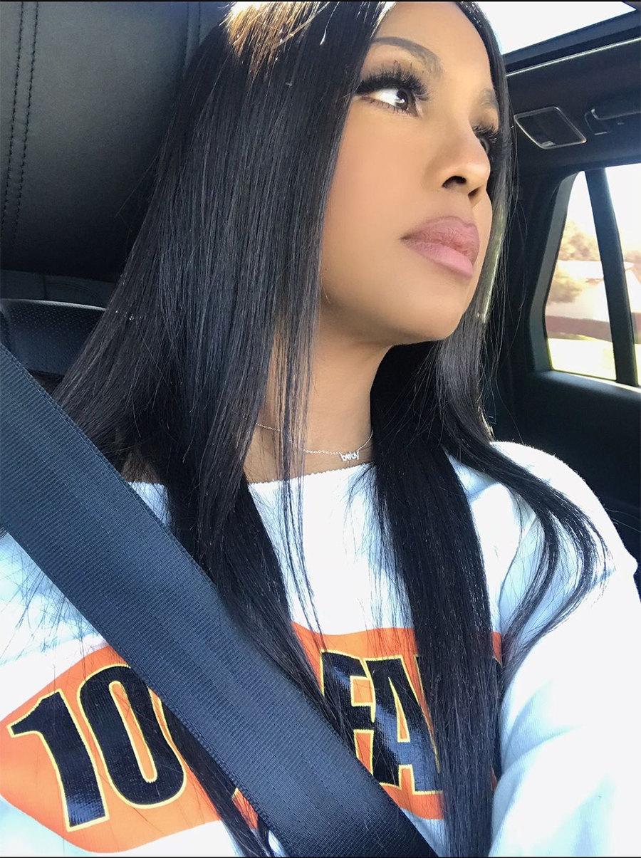 A woman with long straight black hair looks thoughtfully out of a car window while wearing a white shirt with orange and black text and a seatbelt. Sunlight illuminates her face.