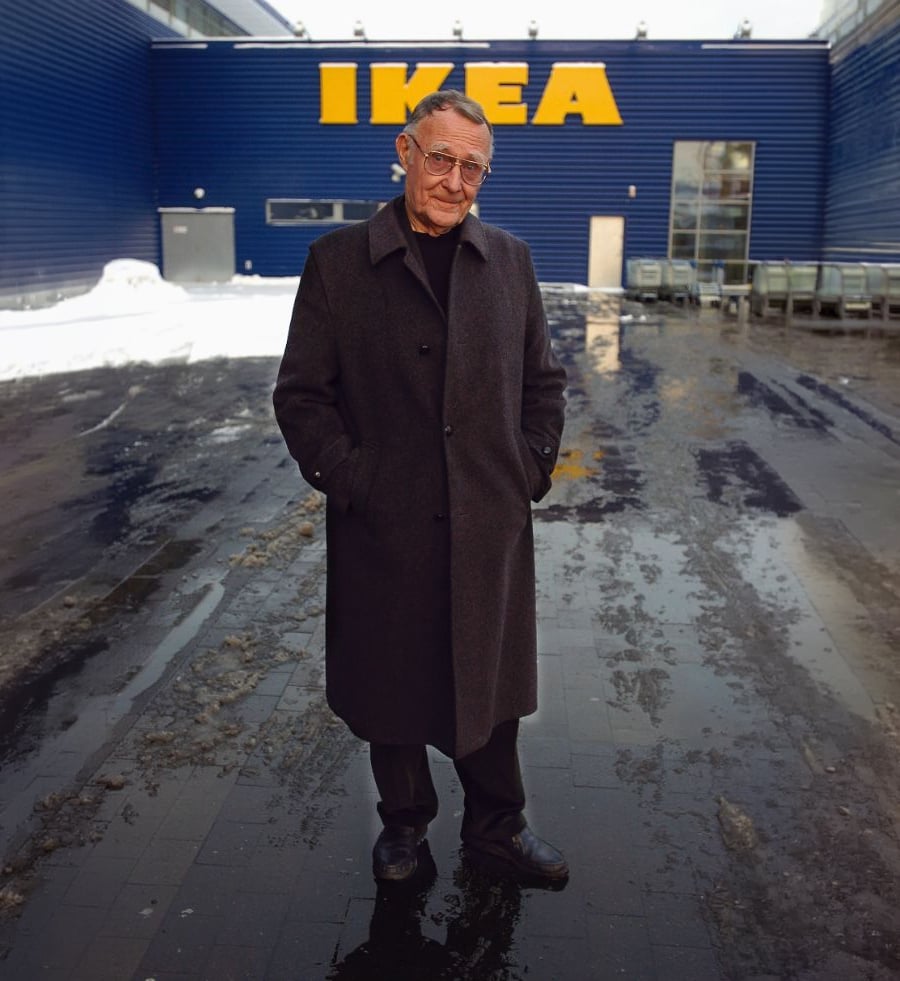 An older man wearing a long dark overcoat stands on a wet pavement outside an IKEA store, with snow piled along the building and shopping carts to the side.