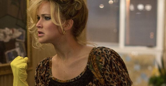 A woman with blonde, messy hair wearing a leopard print top and yellow rubber glove looks to the side with a serious expression in a dimly lit room.