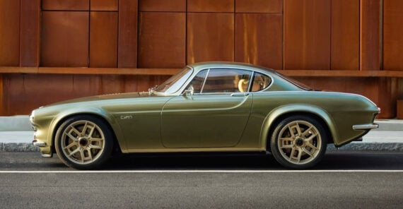 A vintage-style green sports car with gold wheels is parked on a city street in front of a rust-colored metal wall.