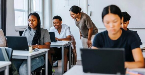A teacher leans over to help a smiling student while others work on laptops in a bright classroom, all focused on their tasks at individual desks.