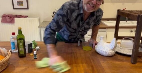 A man wearing glasses and a patterned shirt smiles while cleaning a wooden table with a green cloth in a cozy kitchen. Various items, including a bowl of oranges, a teapot, and bottles, are on the table.