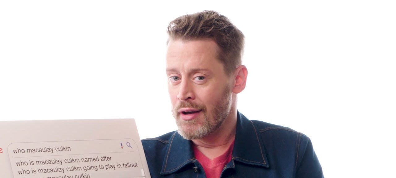 A man with light hair and beard, wearing a denim jacket over a red shirt, holds a board showing Google search suggestions related to "who macaulay culkin" against a white background.