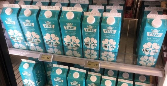 Cartons of TINE Letti Tinemelk milk are neatly arranged on refrigerated shelves in a grocery store. The cartons are blue with white caps and flower designs. Price tags are visible on the shelves below.