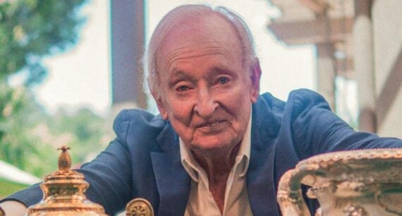 An older man in a blue blazer and white shirt smiles while sitting at a table displaying several large, ornate gold and silver trophies. Trees and a building are visible in the blurred background.
