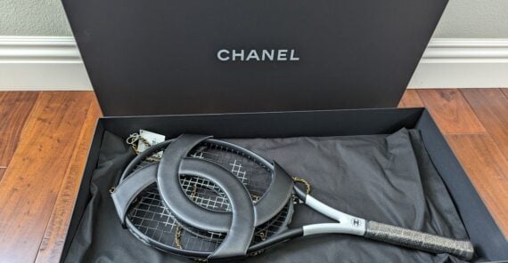 A Chanel-branded tennis racket with a large interlocking "CC" logo is displayed in a black Chanel box lined with fabric, resting on a wooden floor.