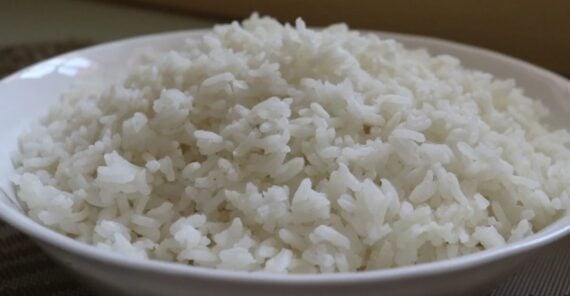 A close-up of a white bowl filled with steamed white rice, placed on a table with a neutral background.