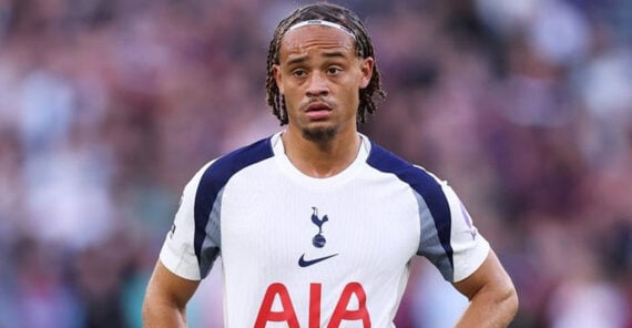 A football player wearing a white Tottenham Hotspur jersey with &ldquo;AIA&rdquo; on the front stands on the field with his hands on his hips, looking ahead with a serious expression.
