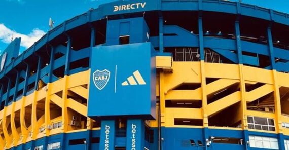 The image shows the exterior of a stadium painted blue and yellow, featuring a large sign with the CABJ crest, an Adidas logo, and advertising banners. The sky is clear and blue above the stadium.