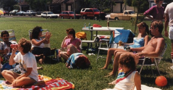 A group of people relax on lawn chairs and blankets in a grassy park, enjoying drinks and chatting. Several cars are parked in the background, and the weather appears sunny and pleasant.