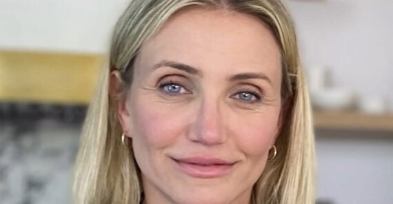 A woman with blonde hair and blue eyes smiles softly at the camera. She is wearing small hoop earrings and natural makeup. The background is softly blurred with light colors.
