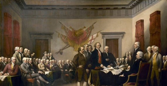 A historic painting depicts a group of men in 18th-century attire gathered in a large room; several stand around a table presenting a document, while others sit and watch, with flags and red drapes in the background.