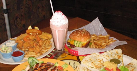 A table filled with fried shrimp, fries, and coleslaw, a strawberry milkshake topped with whipped cream, a cheeseburger with fries, quesadillas with salsa, and a grilled chicken salad with vegetables.