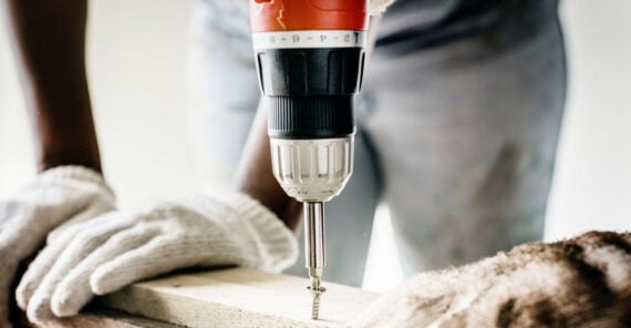 A person wearing white gloves uses a cordless power drill to drive a screw into a piece of light-colored wood. The image is focused on the drill and screw, with the person's body blurred in the background.