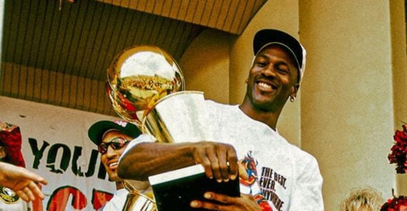 A basketball player in a white shirt and cap smiles while holding a large gold trophy, surrounded by people during a celebration event.
