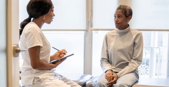 A healthcare professional in white scrubs sits with a notepad, attentively listening to a patient in casual gray clothes who is seated on an exam table in a bright medical office.