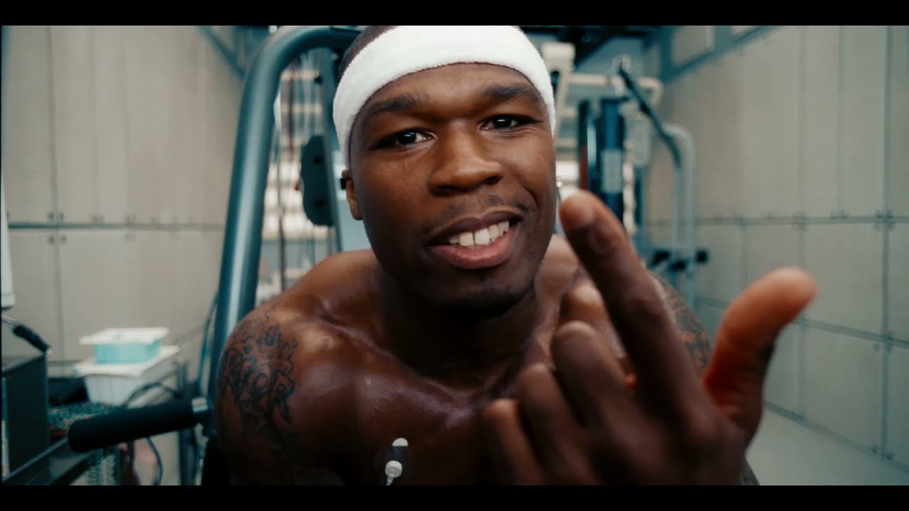 A muscular man with a headband sweats while working out in a gym, looking directly at the camera and pointing with one finger. Exercise equipment is visible in the background.
