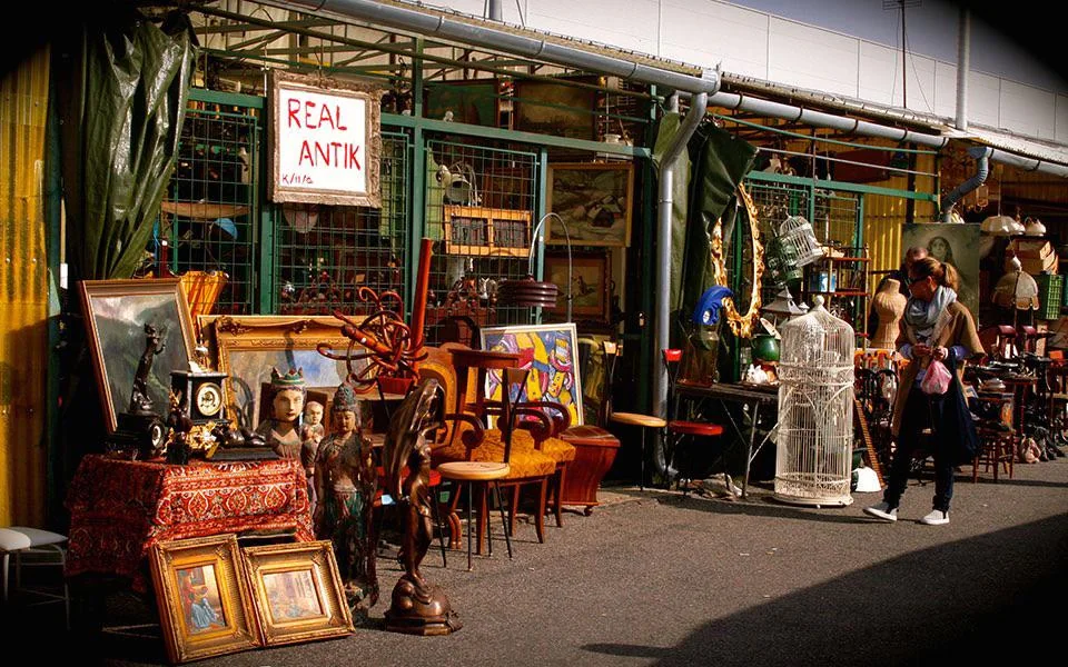 An outdoor antique market stall displays vintage furniture, paintings, clocks, and various collectibles. A handwritten sign reads "REAL ANTIK." Two people walk past the colorful collection on a sunny day.