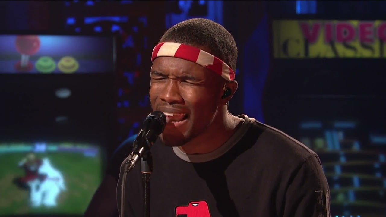 A person wearing a red and white headband sings passionately into a microphone on stage, with retro arcade game machines visible in the background.