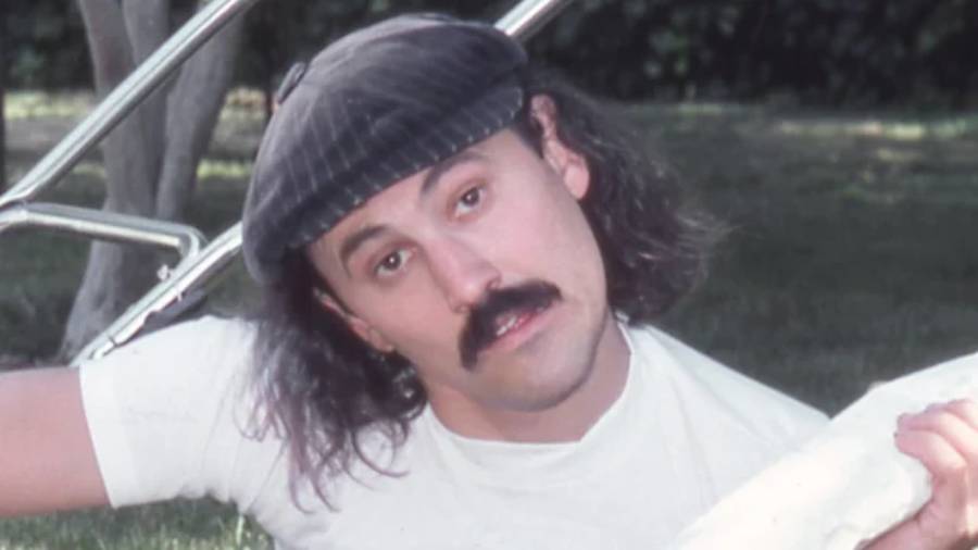 A man with long hair, a mustache, and a flat cap looks toward the camera. He is wearing a white shirt and is outdoors, with grass and a blurred background behind him.