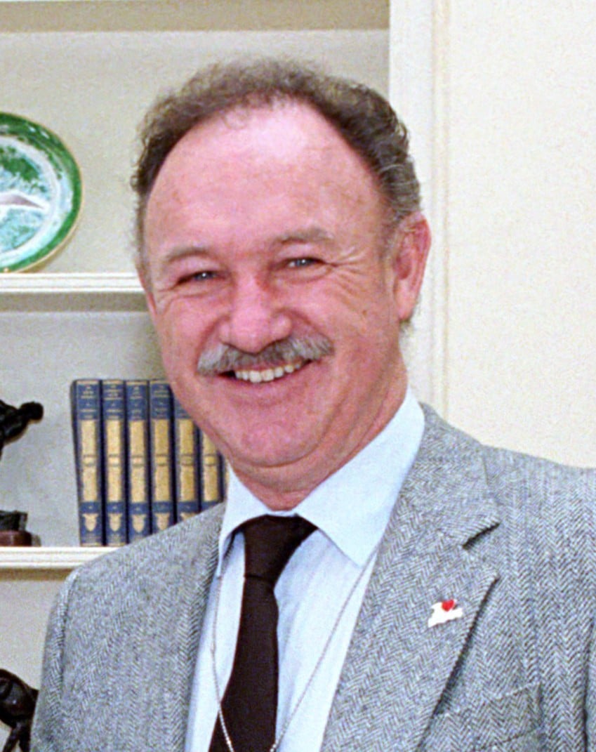 A smiling middle-aged man with a mustache, wearing a light gray blazer, white shirt, and dark tie, stands indoors with shelves containing books and decorative items in the background.