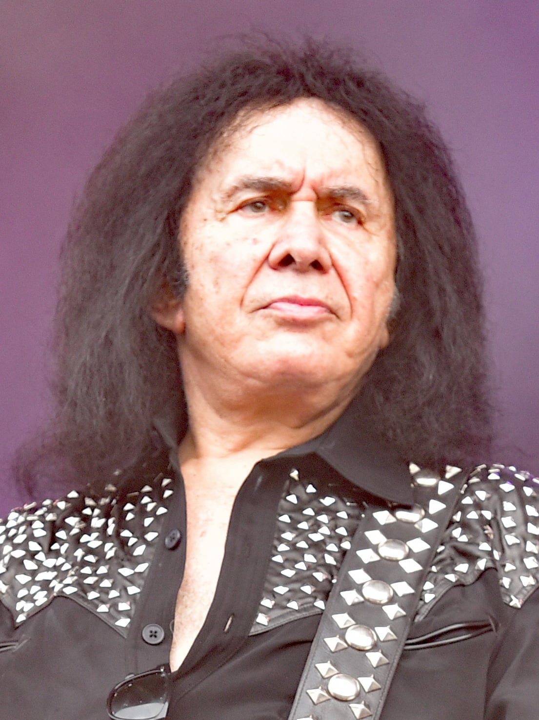 A person with long black hair wearing a black shirt decorated with silver studs and a matching studded strap, looking serious against a purple background.
