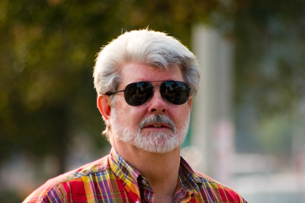 A man with gray hair and a beard, wearing black sunglasses and a red plaid shirt, stands outdoors with blurred greenery in the background.