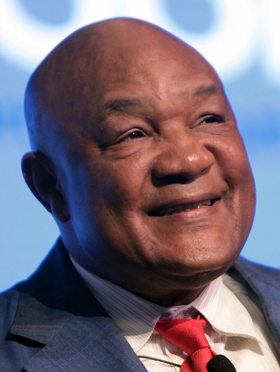 A smiling, bald man in a blue suit, white shirt, and red tie, seen from the shoulders up against a softly blurred blue background.