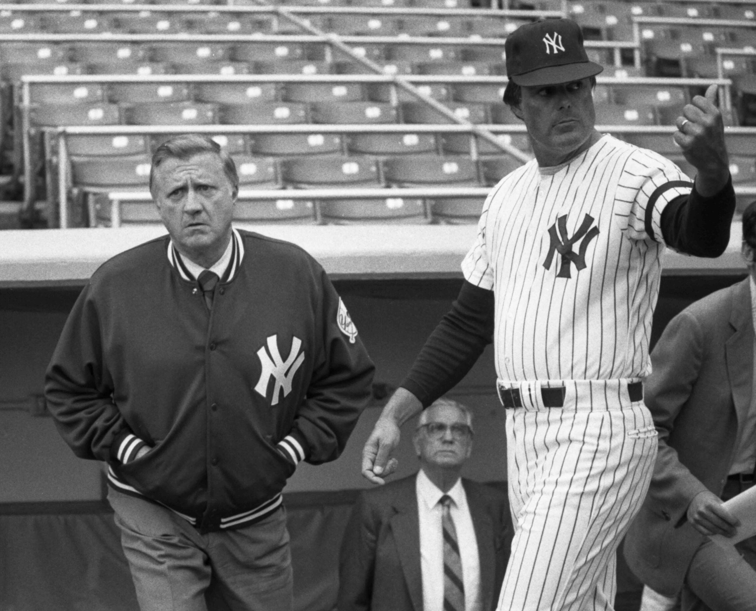 Two men in New York Yankees uniforms stand in a baseball stadium; one wears a jacket and the other, in a striped jersey, gestures with his hand. Empty bleachers and another man in a suit are visible in the background.