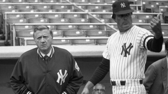 Two men in New York Yankees attire stand in a stadium; one wears a jacket and looks serious, while the other, in a baseball uniform and cap, gestures with his thumb as empty seats fill the background.