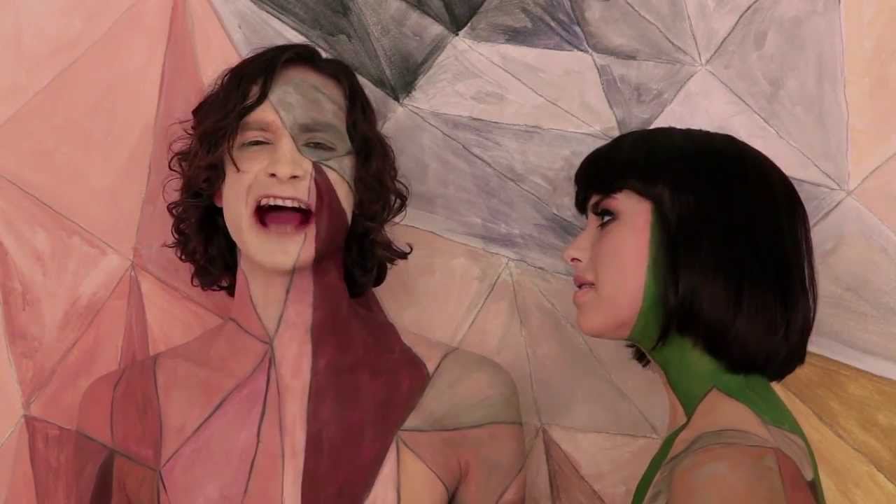 Two people with painted bodies and faces stand in front of a geometric, pastel-colored background that matches their body paint, blending them into the setting. One person faces forward and is singing, while the other looks sideways.