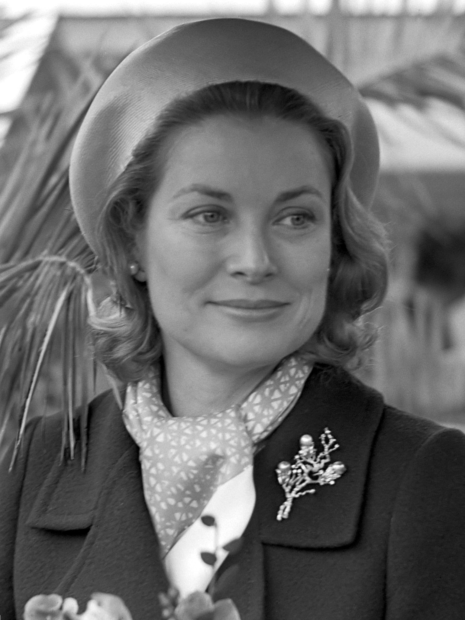 A woman with wavy, shoulder-length hair wears a round hat, a patterned scarf, and a dark coat adorned with a decorative brooch, smiling softly while looking slightly to the side.