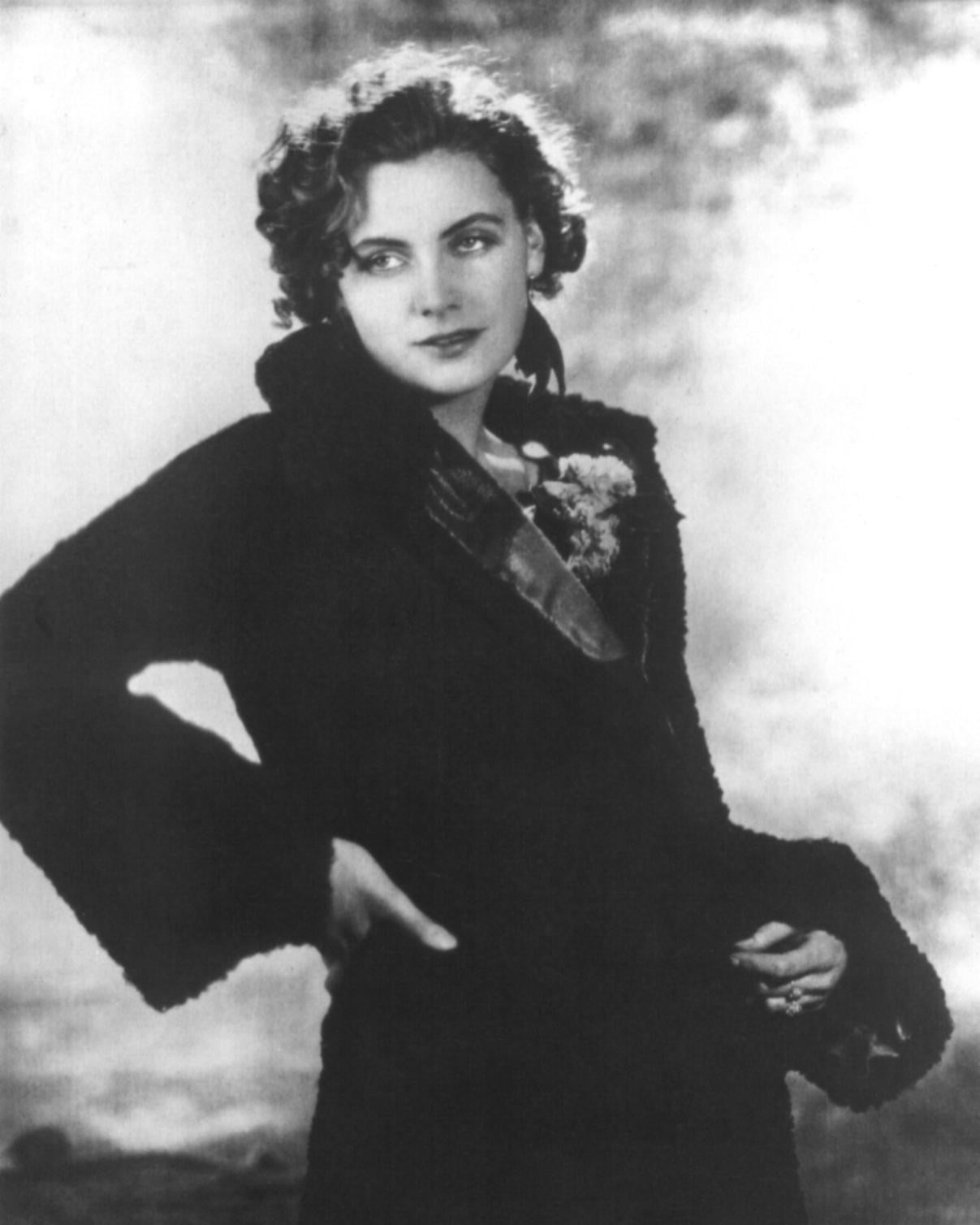 A black-and-white photo of a young woman with curly hair wearing a dark, long coat, looking to the side with a slight smile and her hand resting on her hip against a softly blurred background.