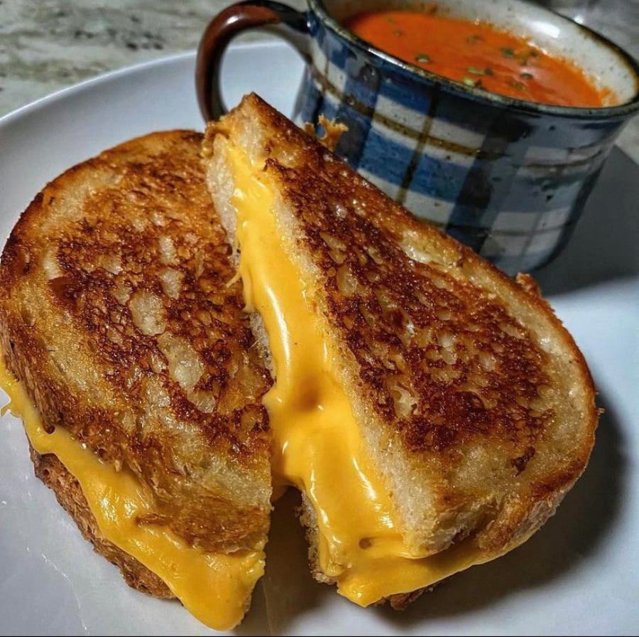 A golden, crispy grilled cheese sandwich with melted cheese oozing out, sliced in half and served on a white plate with a mug of tomato soup in the background.