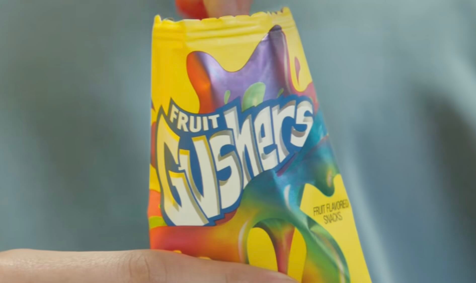 A close-up of a hand holding an opened bright yellow package of Fruit Gushers, a fruit-flavored snack, with colorful graphics and the Gushers logo visible.