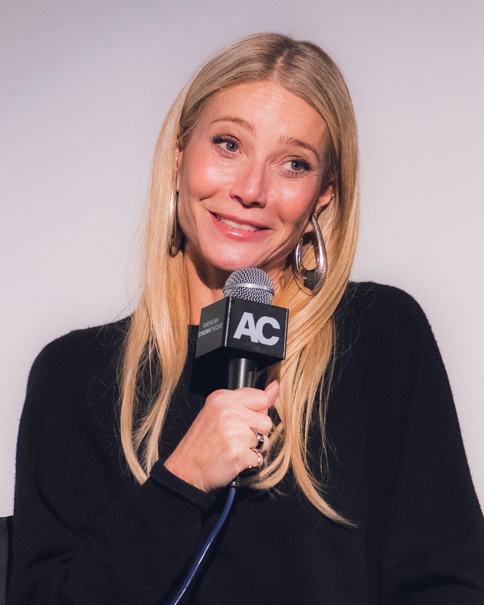A woman with long blonde hair and hoop earrings smiles while holding a microphone labeled "AC." She is wearing a black top and sitting against a plain light background.