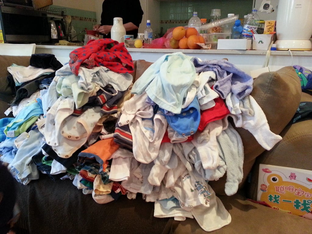 Two large piles of assorted baby clothes are stacked on a couch in front of a cluttered kitchen counter with bottles, fruit, and various items. A person is partially visible behind the counter.