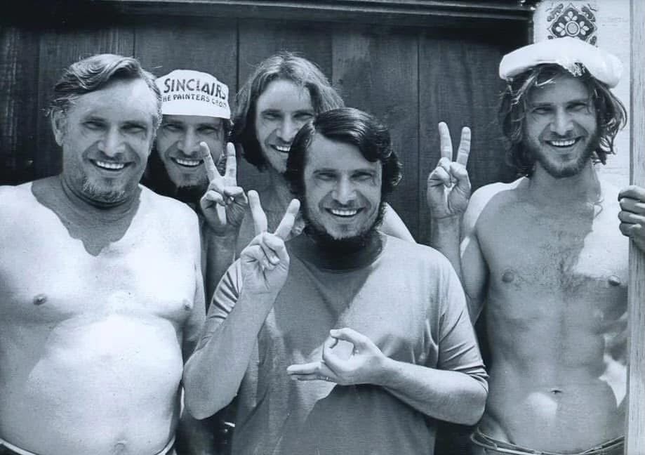 Five smiling men pose together in front of a wooden door, some shirtless, flashing peace signs; one wears a cap and another has a folded sailor hat on his head. The photo is black and white.