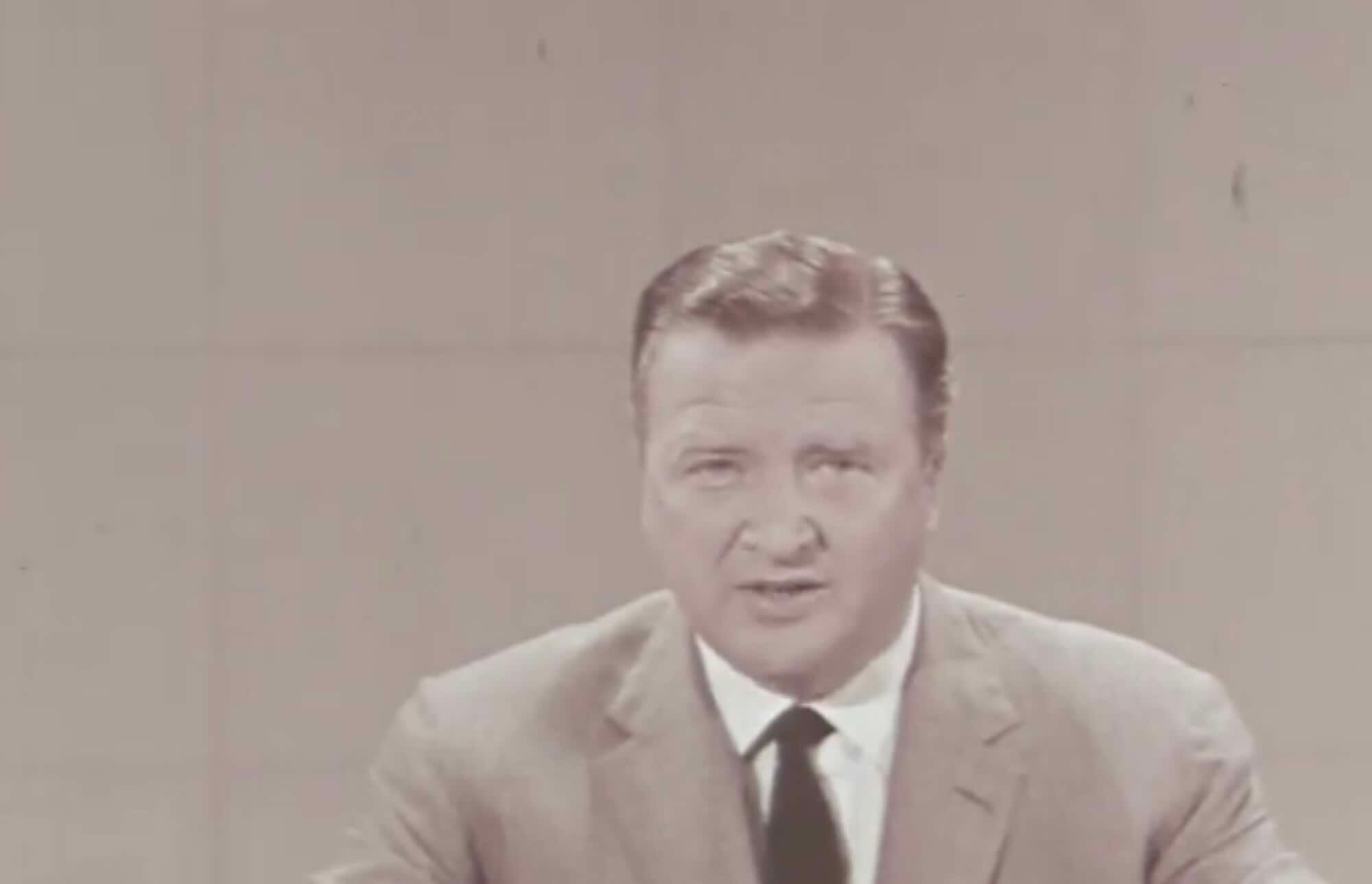 A man in a light-colored suit and dark tie sits at a desk, looking into the camera with a neutral expression, against a plain beige background.