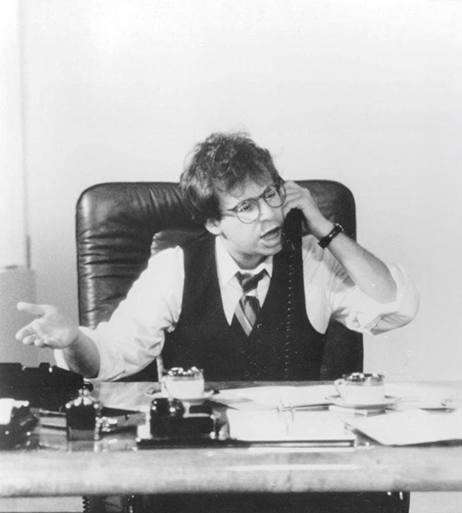 A person with glasses, wearing a vest and tie, sits at a cluttered desk and talks on a corded phone, gesturing with their left hand. The expression appears animated or frustrated.