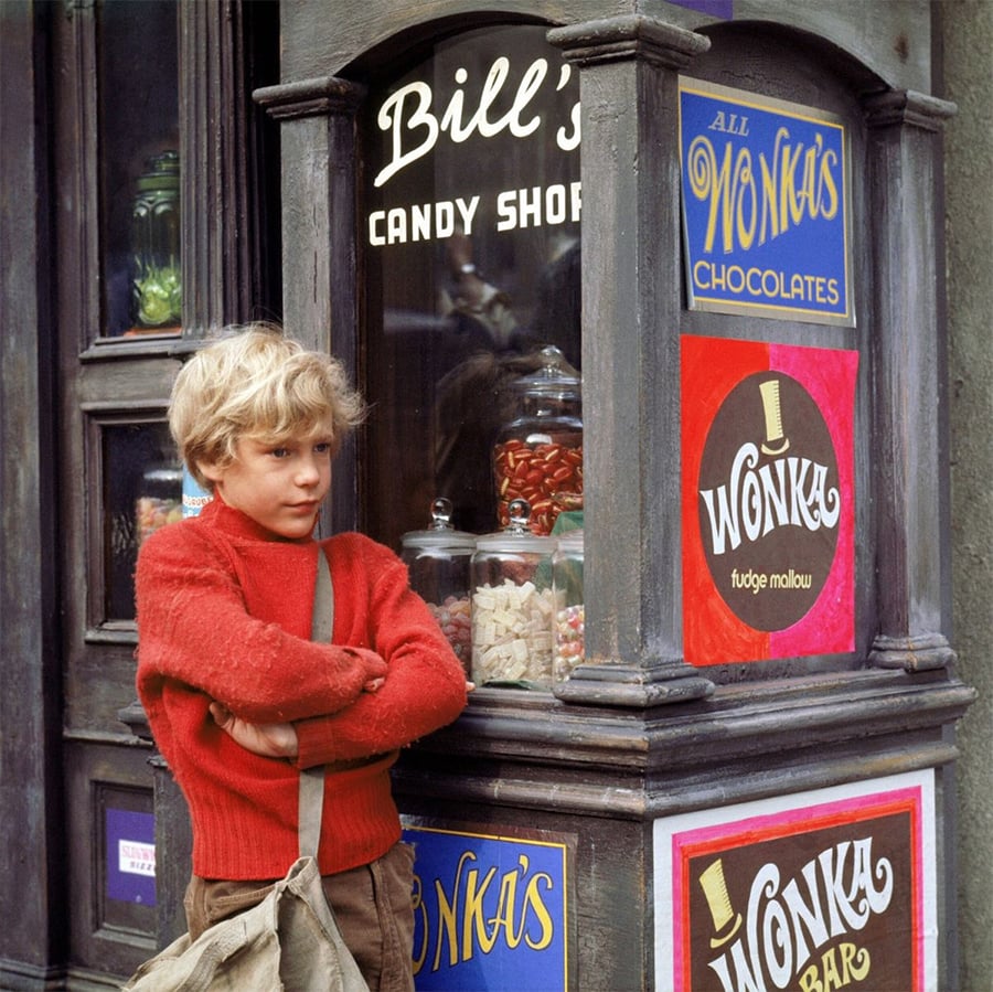 A young boy in a red sweater stands with arms crossed outside a candy shop window displaying jars of sweets and colorful Wonka chocolate posters.