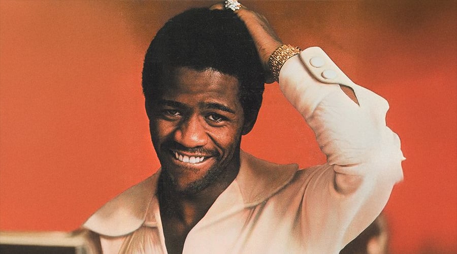 A man smiles at the camera, wearing a white collared shirt and gold jewelry, with one hand touching his head. The background is a solid warm orange.