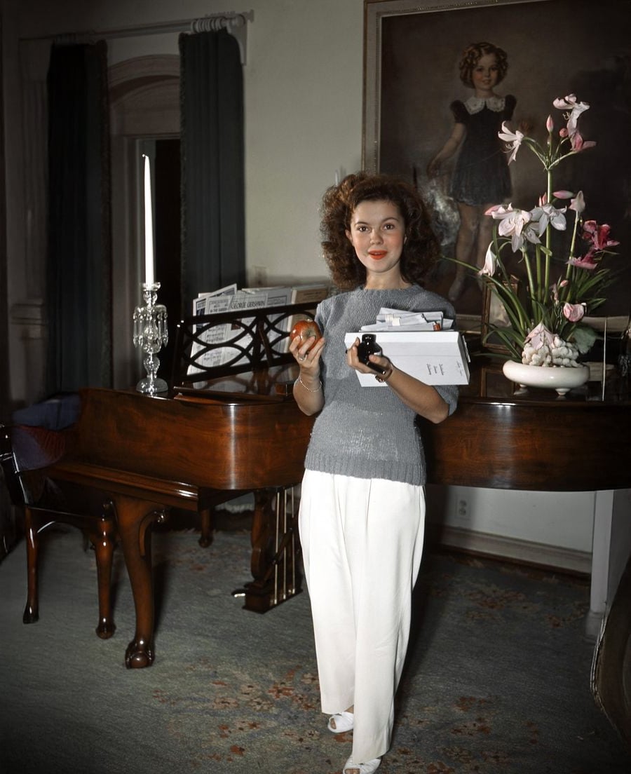 A woman stands indoors, holding an apple and books, in front of a piano with sheet music. A tall candle and a vase of pink flowers sit on the piano, and a large portrait of a young girl hangs on the wall behind her.