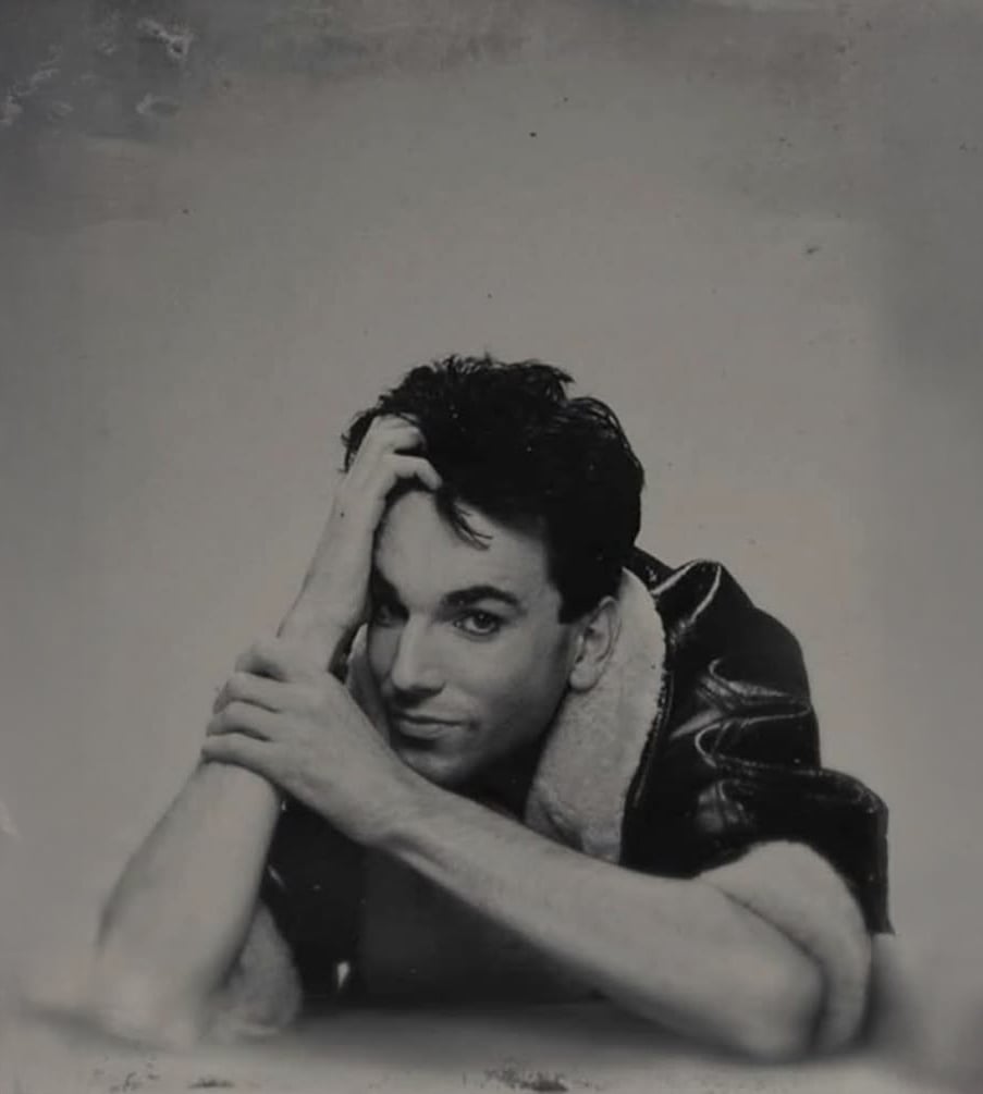 A person with dark hair, wearing a fur-collared jacket, leans forward and rests their head on one hand while looking at the camera with a soft smile, in a black and white portrait.