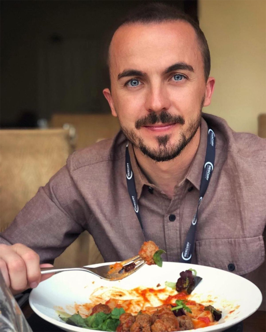 A man with short hair and a trimmed beard, wearing a brown shirt and a lanyard, is sitting at a table and smiling while holding a forkful of food above a plate of meatballs and salad.