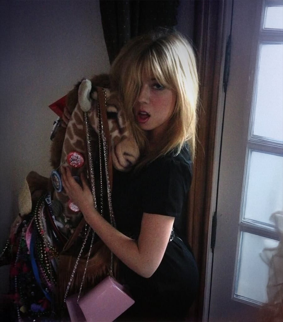 A young woman with blonde hair in a black dress poses indoors, holding a plush giraffe decorated with various pins and necklaces. She stands near a door with glass panels, looking at the camera with her mouth slightly open.