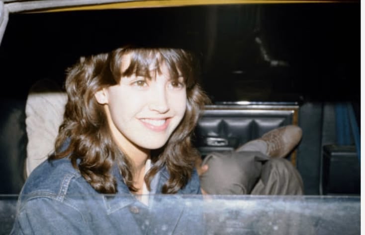 A young woman with wavy brown hair and a denim jacket smiles while sitting in the back seat of a car at night. The interior is dark, and a man's legs are visible next to her.