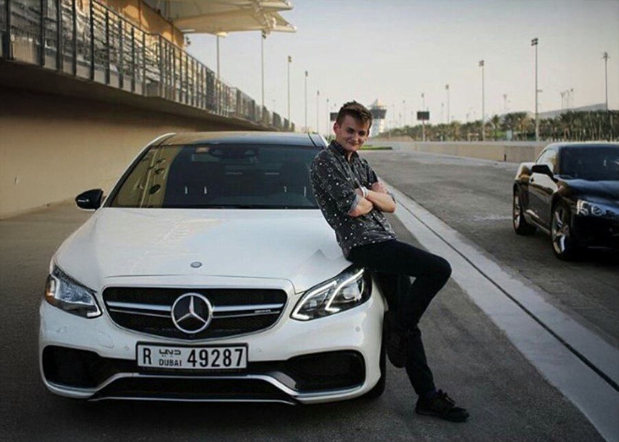 A person leans casually against the front of a white Mercedes-Benz parked on a road, with arms crossed and smiling. The car has a Dubai license plate. Another car is parked nearby, and a fence runs along the background.