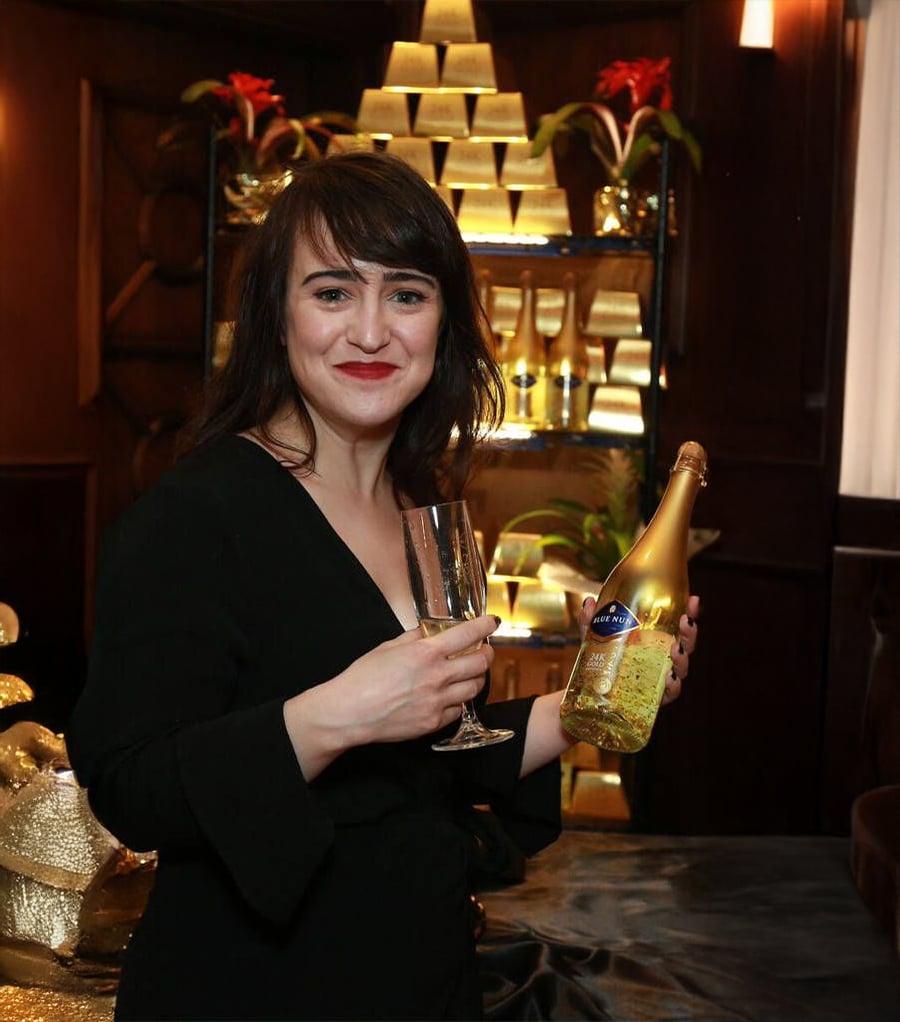 A woman in a black dress smiles while holding a wine glass and a bottle of sparkling wine, standing in front of a pyramid display of gold bars and festive decorations.